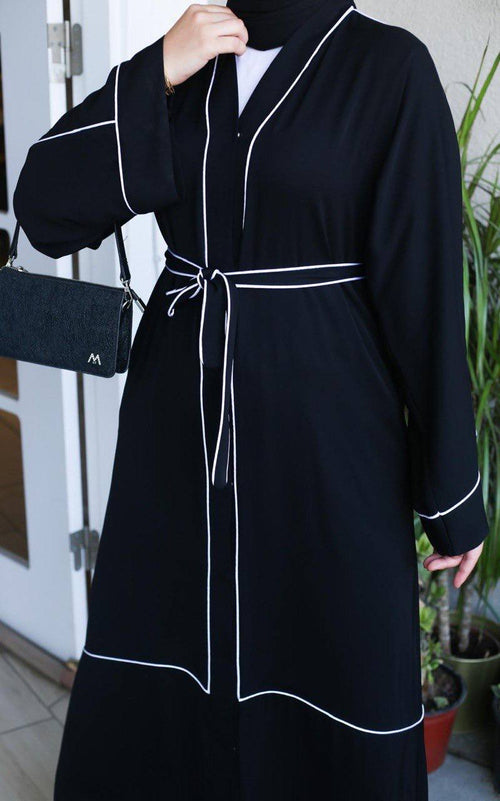 Close-up of black abaya showing white contrast piping along lapel and cuffs with self-tie waist belt detail