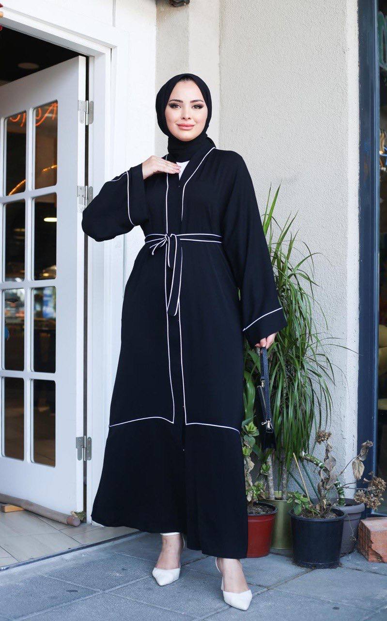 Full-length front view of black abaya with white contrast trim and tiered hem panel, styled with white heels