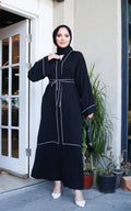 Full-length front view of black abaya with white contrast trim and tiered hem panel, styled with white heels